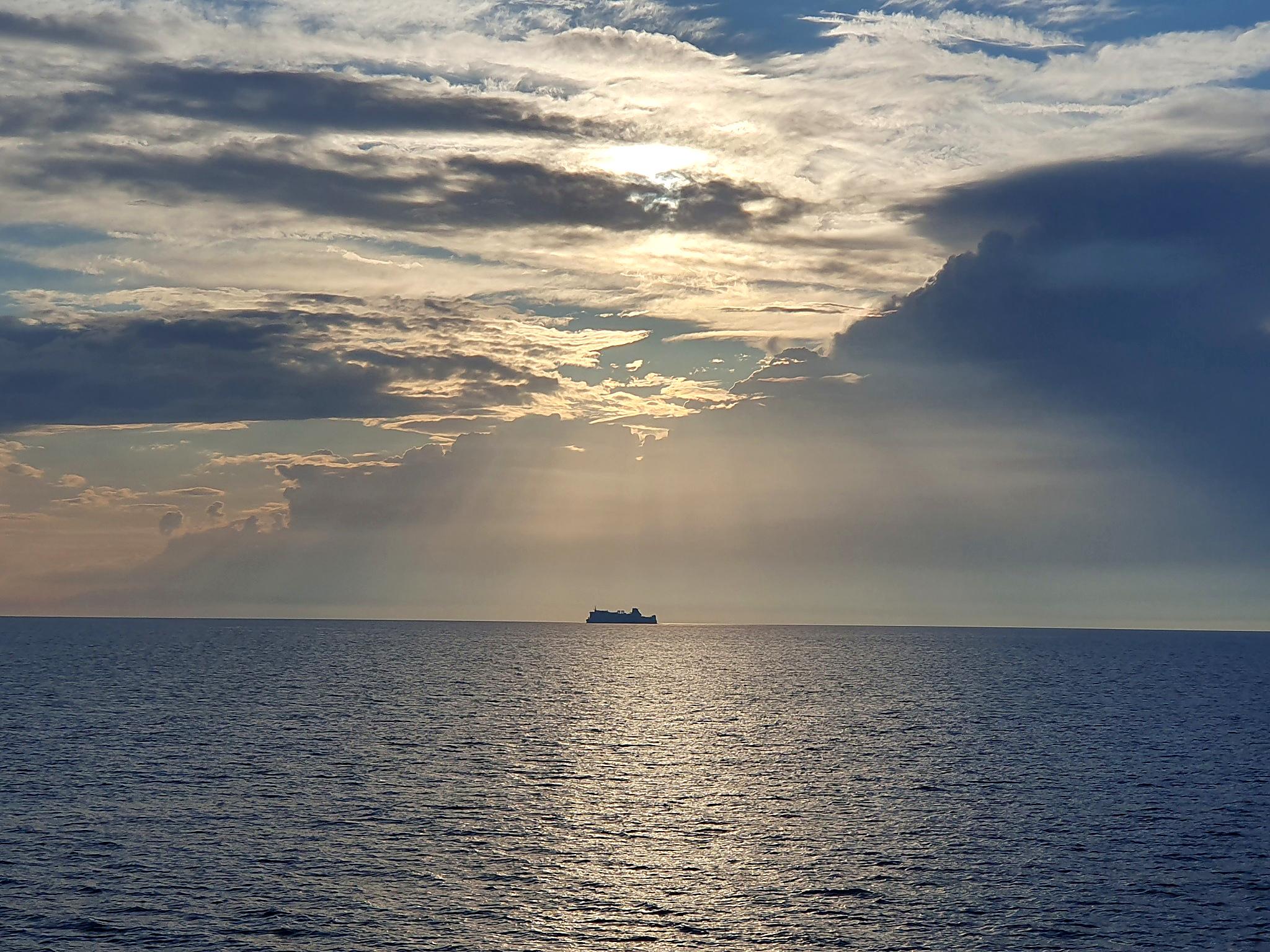 A vessel silhouetted on the horizon under a sunlit, cloud-streaked sky over open sea.