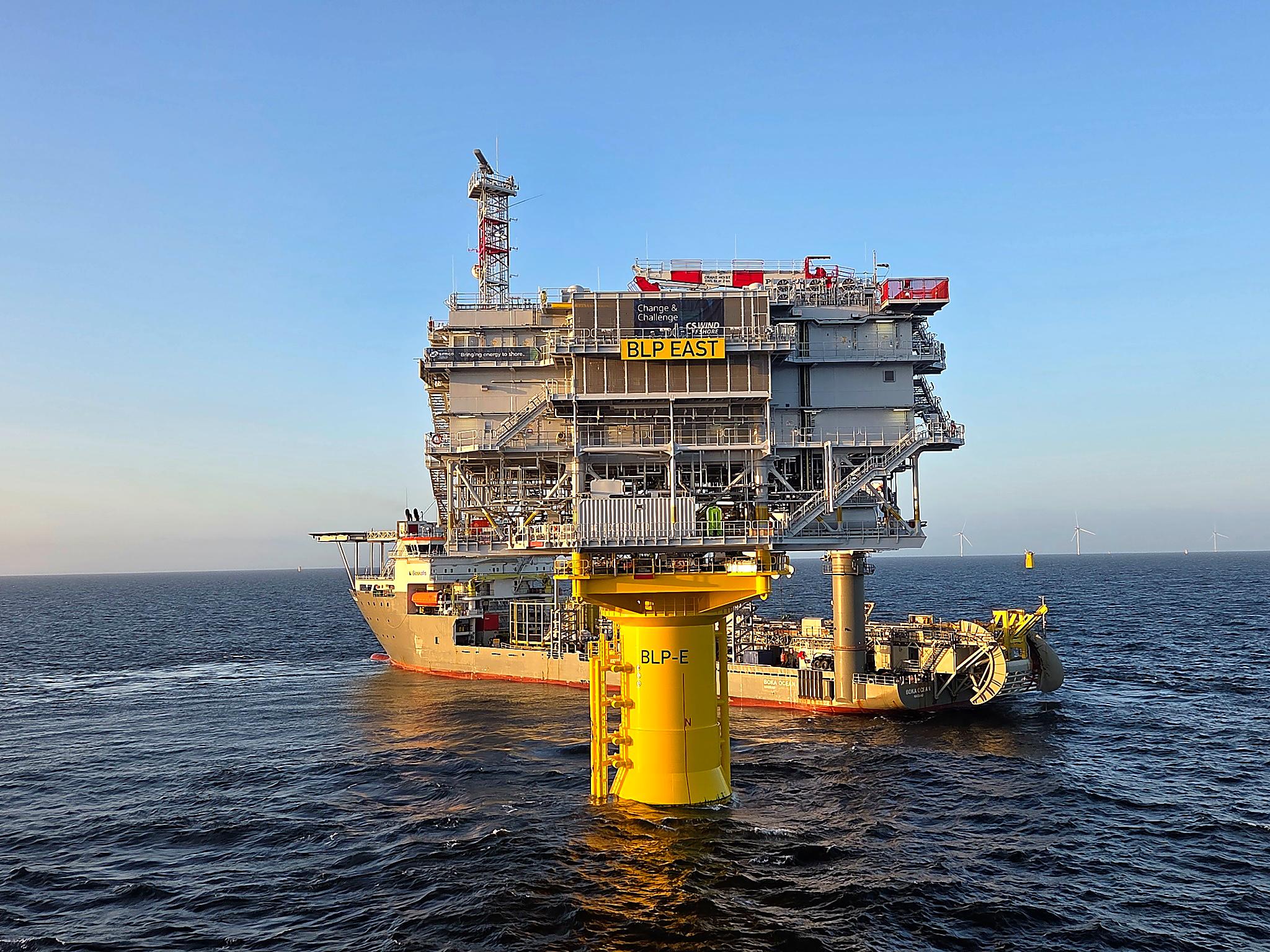 A large offshore energy platform on a yellow jacket foundation, with a support vessel moored beneath it in calm blue water.