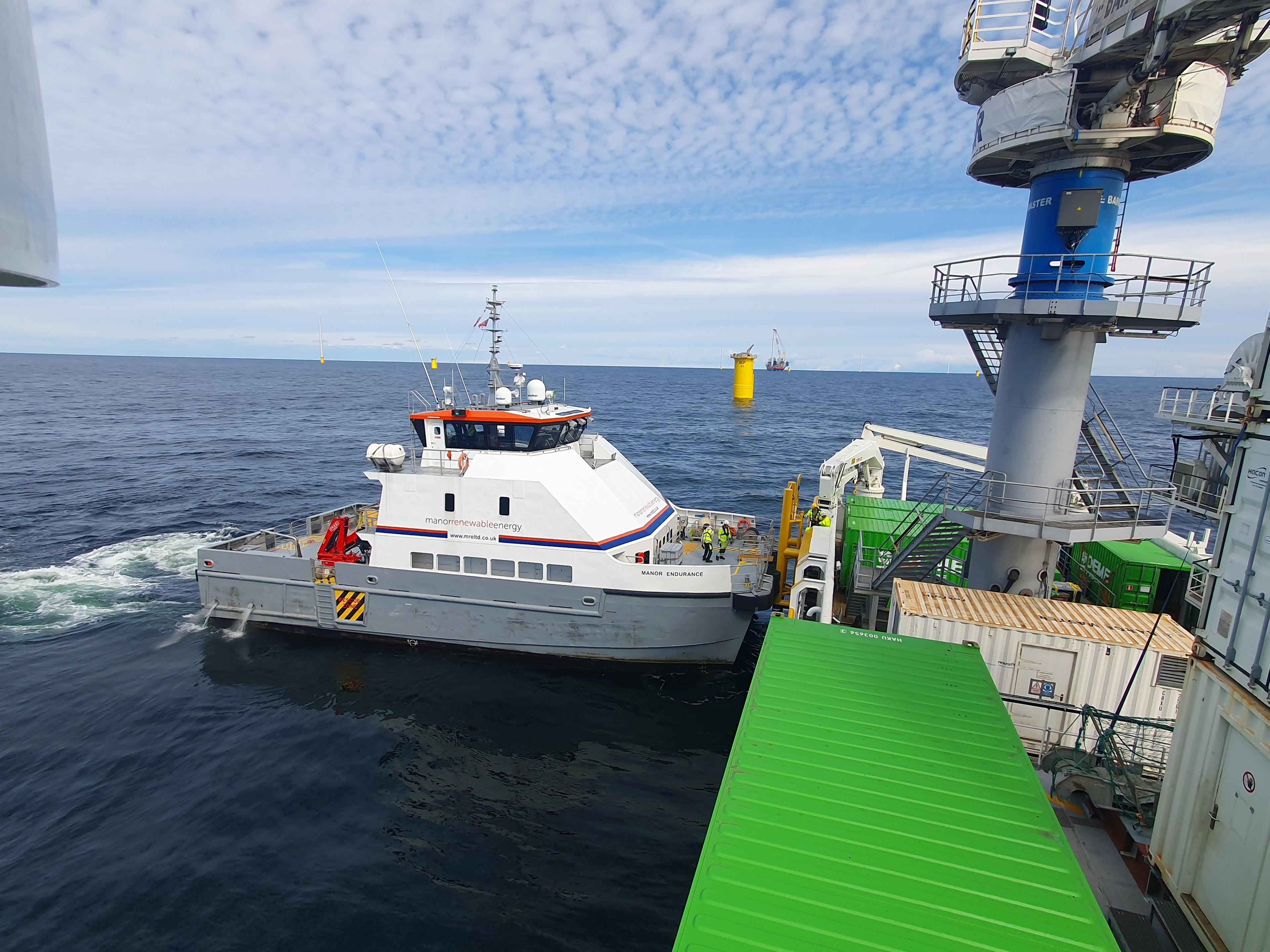 A crew transfer vessel alongside an offshore installation, with crew and cargo containers on the platform deck under a blue sky.