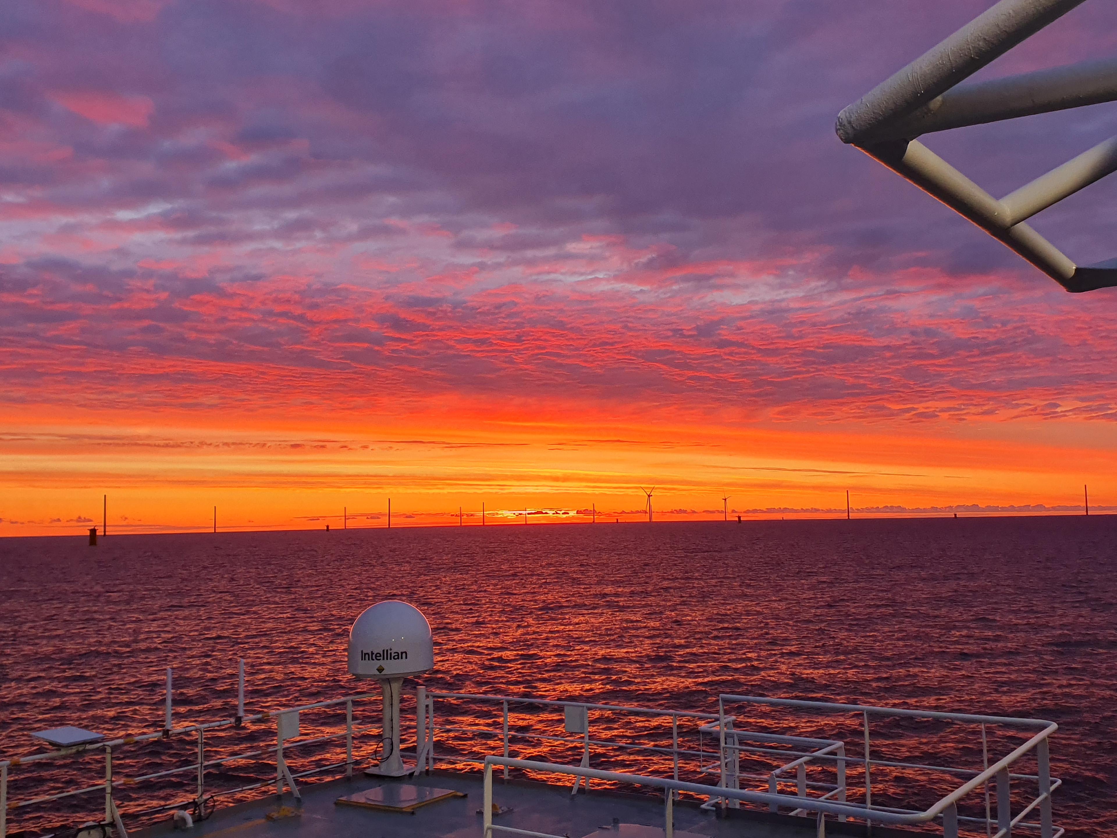 A dramatic red and orange sunset over the sea seen from a vessel deck, with offshore wind turbines lining the horizon and marine equipment in the foreground.