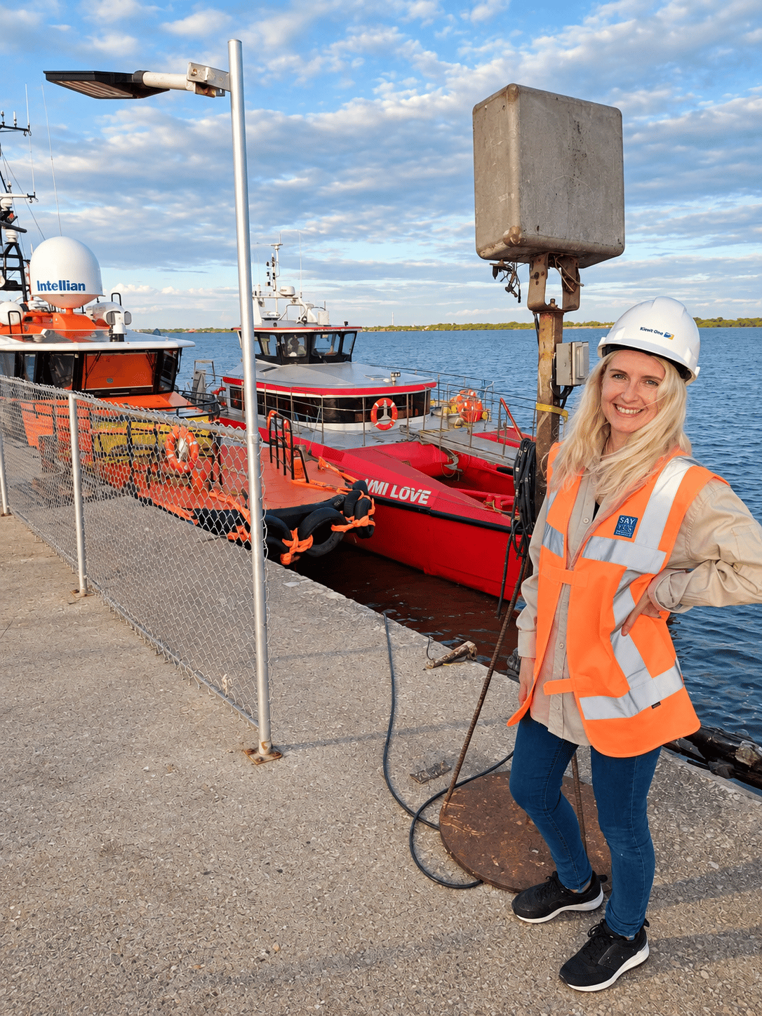 A smiling offshore worker in a hard hat and high-visibility vest standing on a quayside, with crew transfer vessels moored behind her.