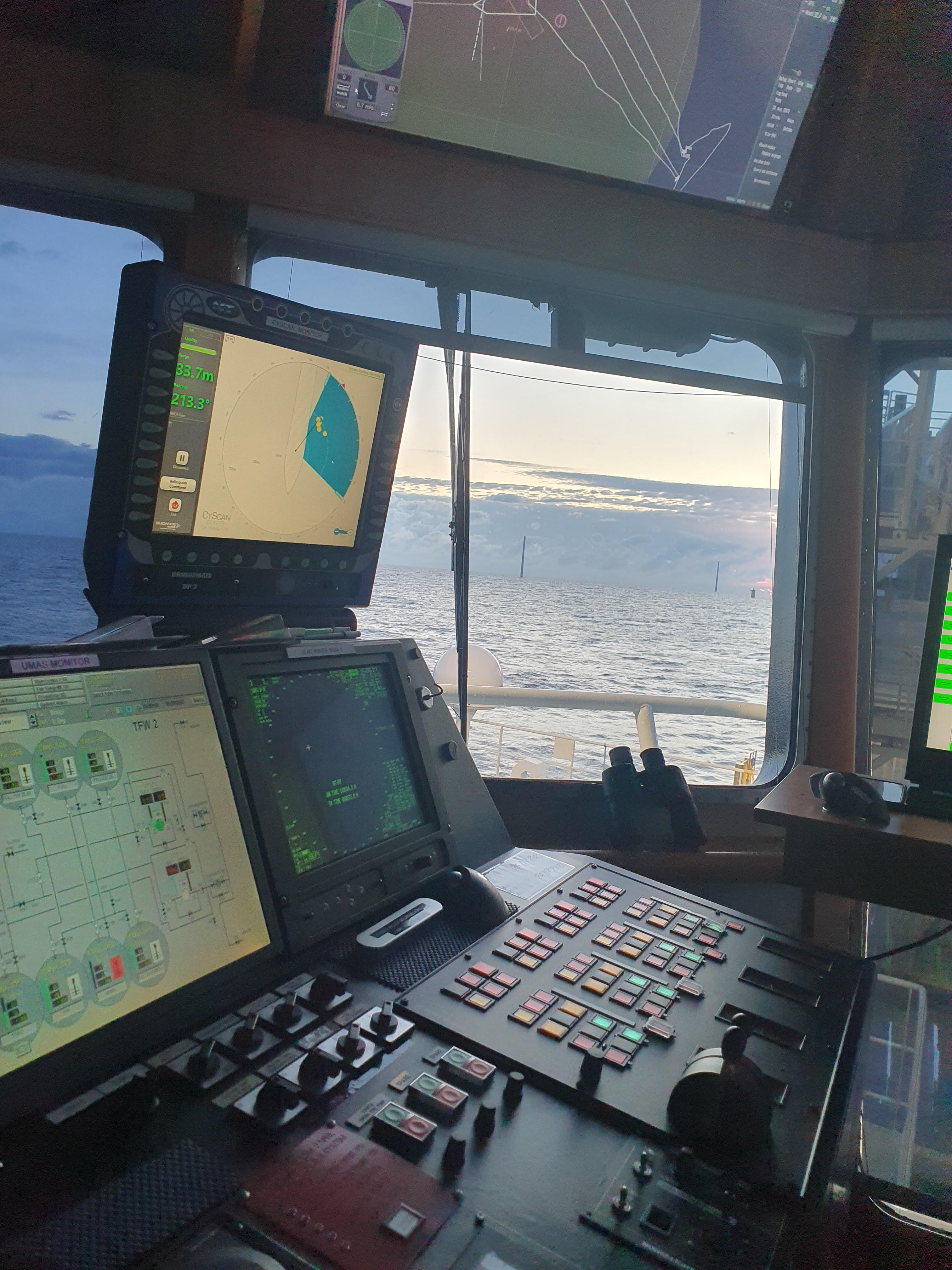 A vessel's bridge control room with navigation displays, radar and engine monitoring screens, looking out across the sea toward an offshore structure.